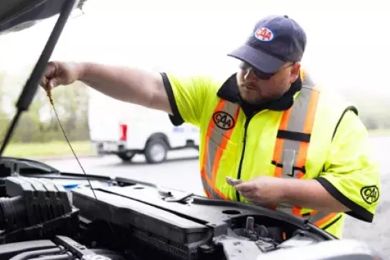 Roadside assistance worker in a bright saftey vest checking under the hood of a car on a highway shoulder with a white truck in the background Roadside assistance worker in a bright saftey vest checking under the hood of a car on a highway shoulder with a white truck in the background