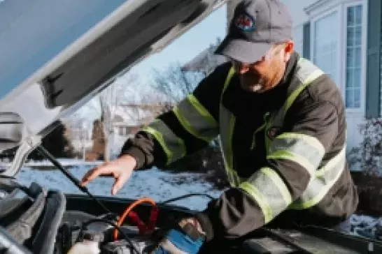 Roadside assistance worker wearing a ball cap and safety gear working under the hood of a vehicle Roadside assistance worker wearing a ball cap and safety gear working under the hood of a vehicle