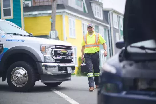 Danny Roadside Tech A roadside assistance worker in a high-visibility yellow shirt and cap walks toward a vehicle with an open hood. A service truck with "Member Service" written on the door is parked nearby. Colorful houses line the street in the background.