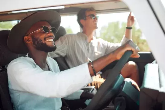 Two men are smiling and enjoying a ride in a convertible. The driver, wearing a large hat and sunglasses, guides the car, while the passenger, also in sunglasses, looks out and enjoys the scenery. Both are dressed in casual, light-colored clothing.
