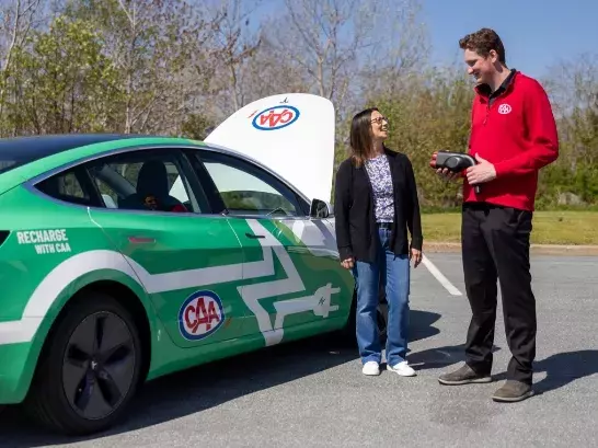 A woman standing next to a CAA worker holding onto an EV charger. Both standing next to a EV with the hood up A woman standing next to a CAA worker holding onto an EV charger. Both standing next to a EV with the hood up