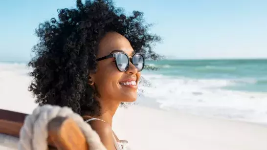 close up shot of a woman smiling with curly hair wearing black sunglasses sitting on a beach chair at the beach